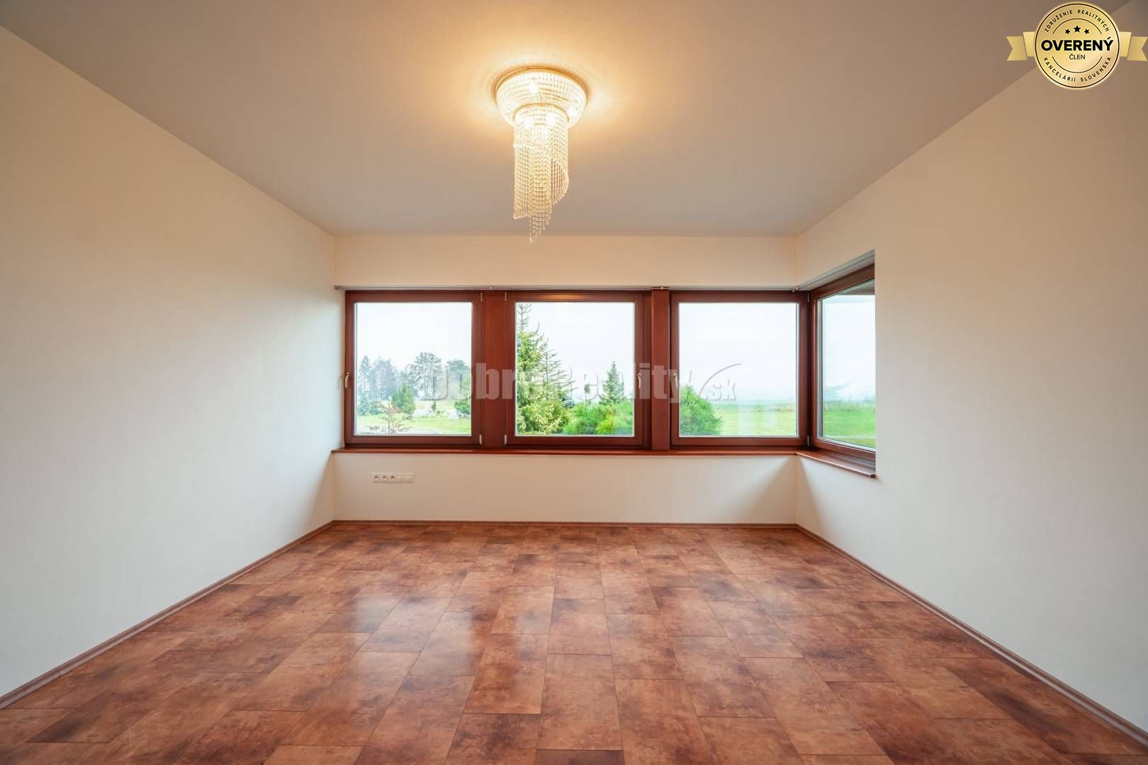 Prázdna miestnosť s veľkými oknami, podlaha s dreveným dekorom, svietidlo na strope.